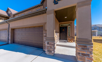 Black front door with decors at the entrance of a house on the side of the garage. House exterior with brown stucco wall and stone veneer walls and brown garage doors on the left.