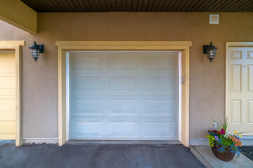 Attached garage exterior with white garage door and wall lights on the side. Garage exterior under balcony with light brown stucco wall and a view of potted flowers and cream front door on the right.
