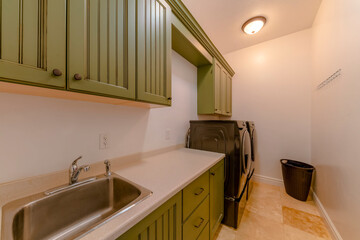 Narrow long laundry room with painted olive green wood cabinets and black washing machines. There is a stainless steel sink on the left below the wall cabinets and black trash can on the right corner.