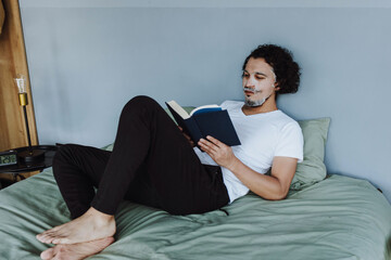 hispanic Man applying exfoliating facial mask treatment and taking care of skin while reading book on bed in loft apartment in Mexico Latin America