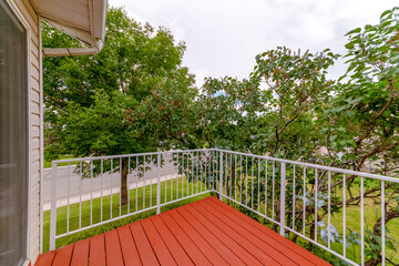 Upper floor deck of a house with red wooden floor and white safety railings. The terrace has a scenic view of the front yard with grassy lawn and lush green trees.