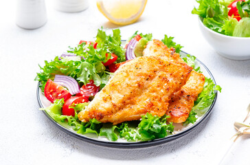 Fried cod fish with salad garnish from lettuce, cherry tomatoes and red onion with sesame seeds, white table background, top view