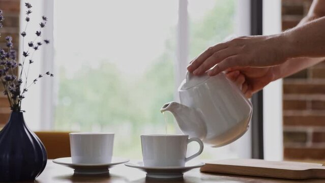 Hand Is Pouring Hot Drink From Teapot Into Two Tea Cups, Mugs Indoors. Relaxing Time At Home Or In Cafe Or Restaurant. Waiter Serving Customers. Business With Good Service. High Quality FullHD Footage