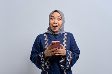 Excited young Asian Muslim woman using mobile phone, receiving good news isolated over white background