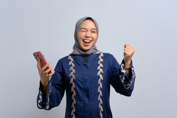 Excited young Asian Muslim woman using mobile phone and celebrating success, getting good news isolated over white background