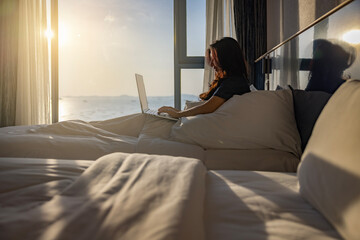 Happy fresh relaxed asian woman resting sitting on bed in hotel room with two beds using laptop enjoying good morning after wakeup in cozy room with soft light from window. 