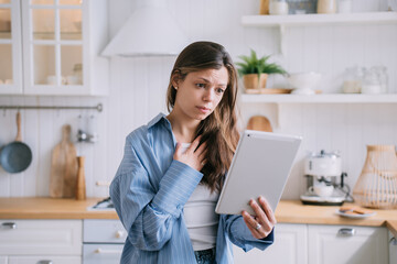 Confused pretty caucasian woman makes video call using tablet touches chest feels frustration unhappy by received news. Dark haired Italian housewife in blue shirt talks with parents at kitchen. Grief