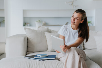 Young creative interior designer sitting on cozy couch holds tablet toothy smiles looks aside. Beautiful american student girl remote learning home. Purposeful female entrepreneur planning business.