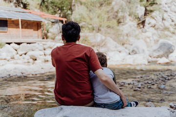 Rearview of school boy and his dad with yellow backpack sits on a riverside rock in canyon with mountain cliffs in the background. Kid child and father taking a rest on a boulder near mountain river.