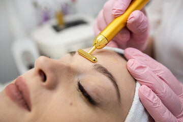 Naklejka premium woman receive pulse face massaging at spa, vibrating golden facial massager on female face
