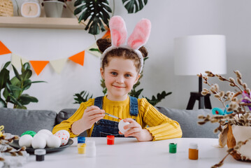 Portrait of sweet little girl child wearing pink fluffy bunny ears sitting at table and painting eggs while preparing for Easter celebration, happy cute kid looking at camera. Spring holiday concept