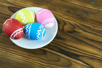 Multicolored Easter eggs on white saucer, wooden background. Happy Easter greeting background with space for text.