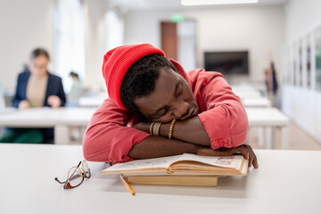 Lazy African male student falling asleep during study, feeling drowsy while studying in library, selective focus. Black guy sitting at desk sleeping on textbooks, napping in classroom. College burnout