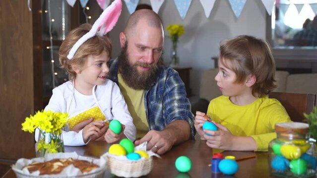 Easter Family traditions. Father and two caucasian happy children with bunny ears dye and decorate eggs with paints for holidays while sitting together at home table. Kids embrace and smile in cozy.