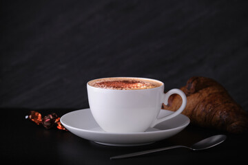 Italian breakfast on a black background. Cappuccino with croissant on a black background. White cup with cappuccino on a dark background.