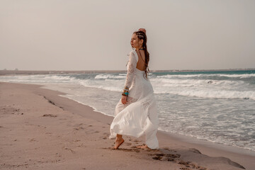Fototapeta na wymiar Model in boho style in a white long dress and silver jewelry on the beach. Her hair is braided, and there are many bracelets on her arms.