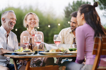Family friends having dinner around table in a garden in summer, drinking wine, talking and laughing in outdoor restaurant at night - obrazy, fototapety, plakaty