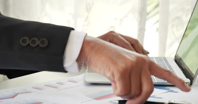 Closeup businessman hands working use laptop finance business document reading report paperwork spreadsheet at office desk. Man hands type keyboard write on office note check financial graph chart
