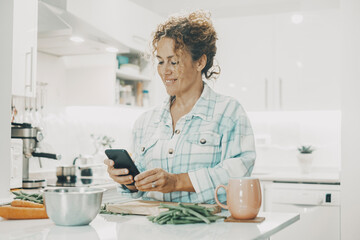 Happy woman at home preparing food lunch and watching mobile phone to follow instructions or tutorial or new idea recipe. Modern online female people chatting alone. Using cell in indoor leisure