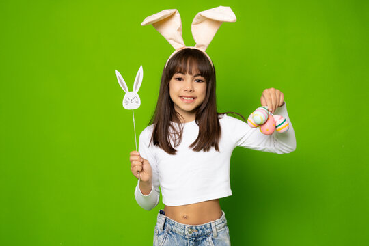 Photo of joyful brunette girl in toy rabbit ears smiling and holding colorful eggs isolated over green background