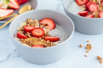 Chia pudding with homemade coconut granola, peanut butter and strawberries in a gray bowl, marble background. Healthy diet, detox, summer recipe.