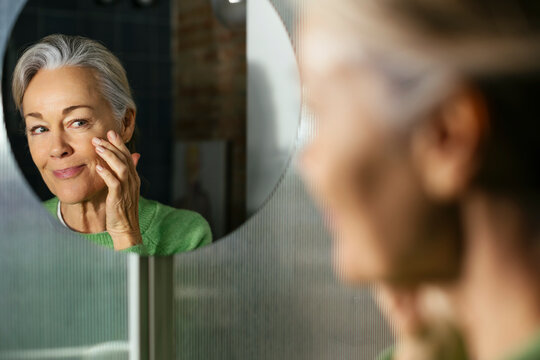 Mature Woman Applying Moisturizer On Face Looking In Mirror