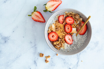 Chia pudding with homemade coconut granola, peanut butter and strawberries in a gray bowl, marble background. Healthy diet, detox, summer recipe.