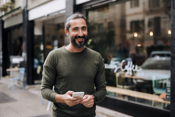 Happy mature man with smart phone standing on footpath