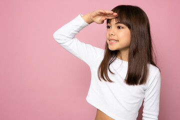 Young beautiful child girl wearing white t-shirt standing over isolated pink background very happy and smiling looking far away with hand over head. Searching concept.