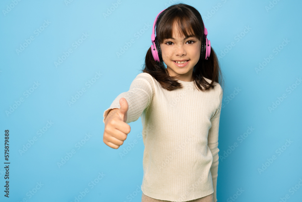 Wall mural Photo of sweet funny young girl wear earphones with thumb up enjoying music isolated blue color background