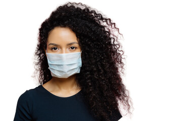 Serious dark skinned young woman being on self isolation at home, wears protective medical mask,...