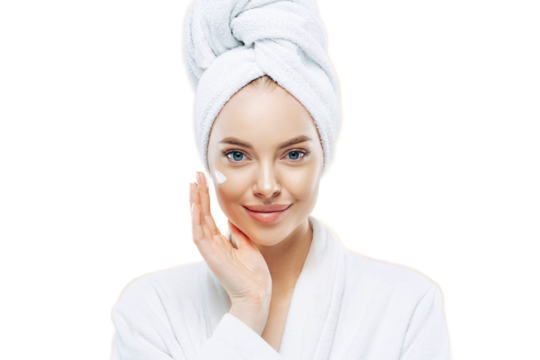 Young confident Caucasian woman applies face cream, enjoys new anti wrinkle cosmetic product, prevents sign of skin aging, wears minimal makeup, dressed in bath robe, isolated on white wall.