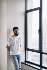 Fototapeta na wymiar A guy with a beard in white T-shirts and blue jeans stands by the window in a new apartment and rejoices in happiness.