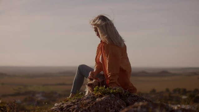 A young woman in an orange jacket sits on a mountain peak at sunset, holding her small dog in her arms on a windy day. happy watching the sunset with a beautiful view