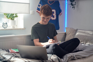 Caucasian teenager boy sitting on bed and learning using laptop