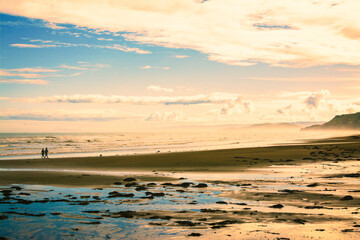 Naklejka premium Active couple walking their pet dog along a vast sand beach at sunset. Pacific Ocean coastline near Gisborne, New Zealand
