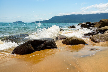 Blue sea, mountains in the background and waves breaking on the sand stones in Ilhabela, São Paulo