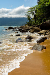Side view of a quiet beach, foam from gentle waves on the rocks and mountains covered in clouds in the background. Ilhabela, Sao Paulo