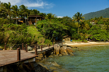 Quiet corner of the island seen from the small wooden pier. Small stretch of beach with yellow sand, surrounded by mountains, palm trees and Atlantic forest. Ilhabela, Sao Paulo