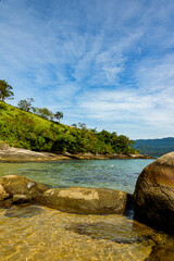Vertical image of a deserted beach with clear water and blue sea surrounded by rocks. In the background mountains covered by tropical forest and sky covered with clouds. Ilhabela, Sao Paulo