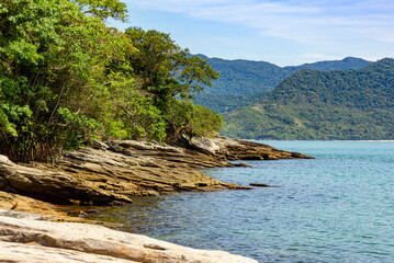 View of the island's rocky corner, surrounded by mountains, tropical forest and calm blue sea in Ilhabela, Sao Paulo