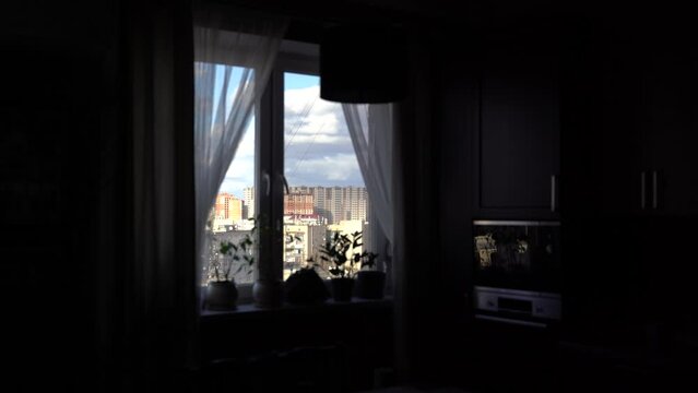 A dark, gloomy room in a high-rise building with a large window on the windowsill there are many different colors. There is a big city outside the window and the warm sun is shining.