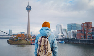 Fototapeta na wymiar autumn or winter travel to Dusseldorf, Germany. young Asian tourist or student in blue jacket and yellow hat ( symbol of Ukraine) walks through sights of European city. beautiful view in the Media Bay