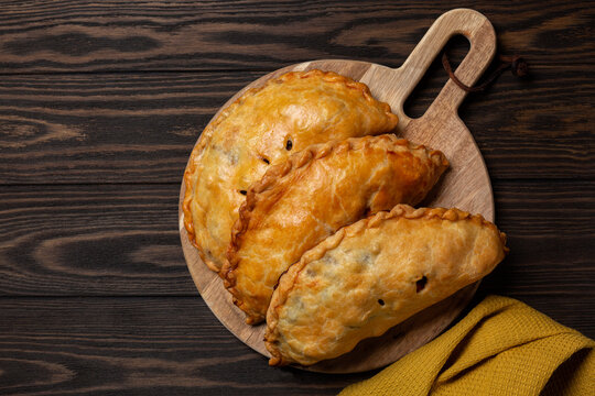 Flat la of baked meat turnovers or pies, or empanadas, or cornish pasty with filling, beef, carrot, and potato on a dark wooden board.