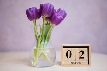 Hello spring! Wooden cubic calendar with date of 2 March surrounded with bouquet of purple tulips with green leaves in glass vase.