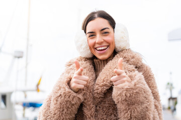Young woman wearing winter muffs at outdoors pointing to the front and smiling