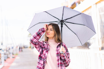 Young pretty Romanian woman holding an umbrella at outdoors having doubts and with confuse face expression