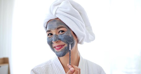 Asian woman applying grey mask on face smiling at camera. Spa beauty procedures at home for girl....