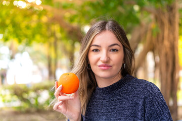 Young pretty Romanian woman holding an orange at outdoors with sad expression