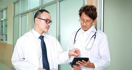 Two Asian doctors talking and discussing patient diagnosis data hold digital tablet computer technology in the hospital hallway